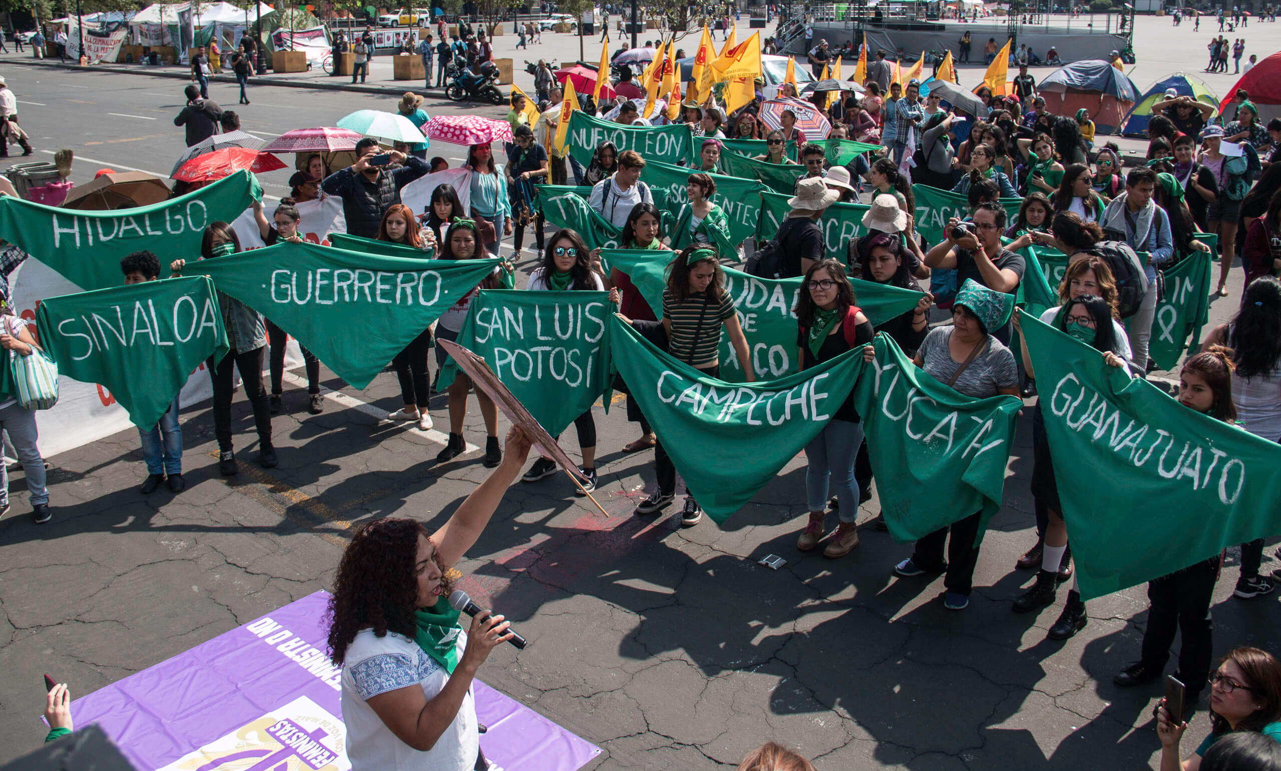 Fotografía en plano general de un grupo de personas que sostienen pañuelos verdes aborteros en tamaño grande. Cada uno tiene el nombre de las entidades de la República mexicana.