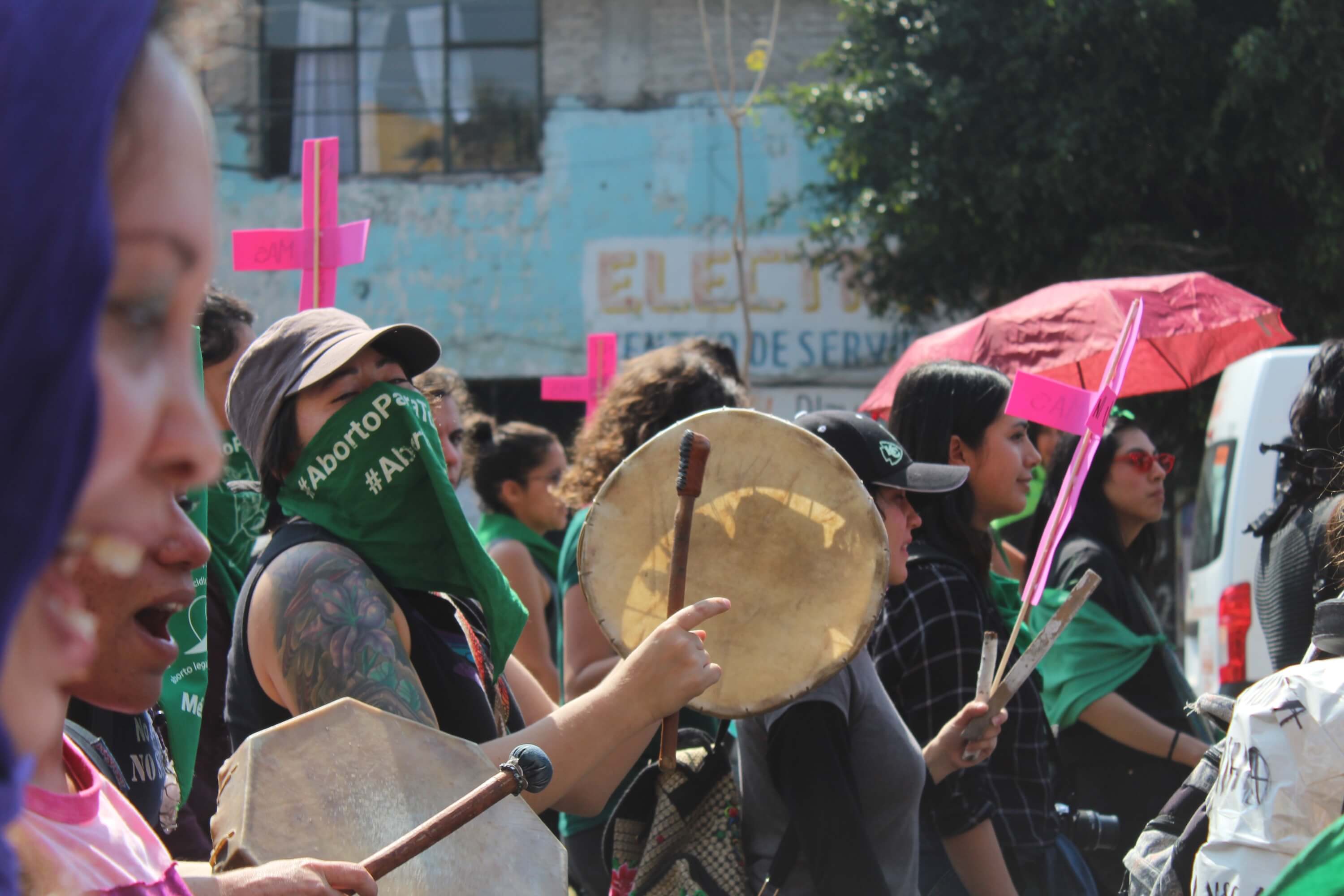Fotografía en plano medio de una mujer que tiene el rostro cubierto con un pañuelo verde abortero. Está en una manifestación tocando un tambor.
