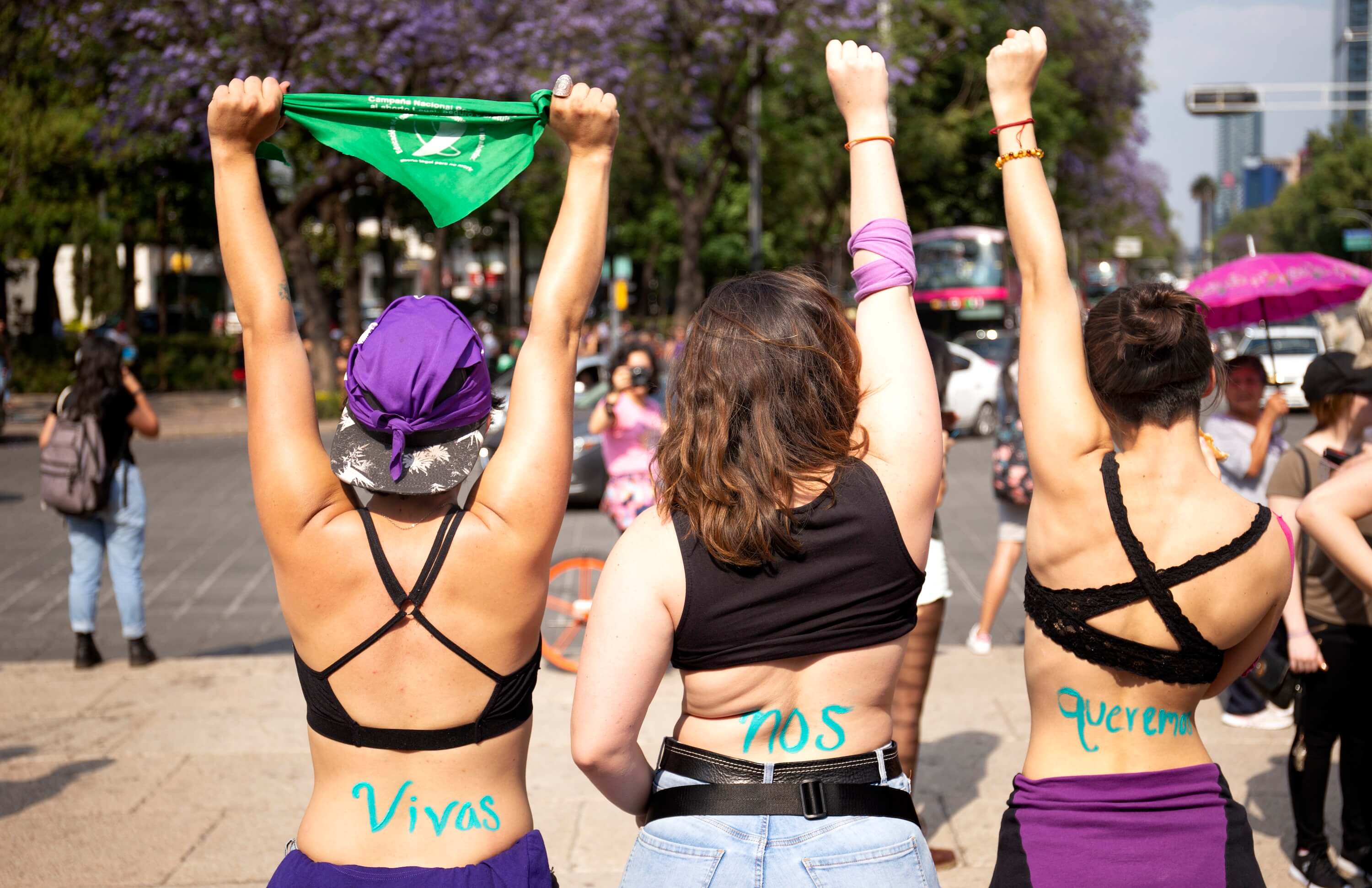 Fotografía en plano medio de tres mujeres que son retratadas de espaldas. Dos de ellas alzan su puño mientras que otra sostiene un pañuelo verde abortero.