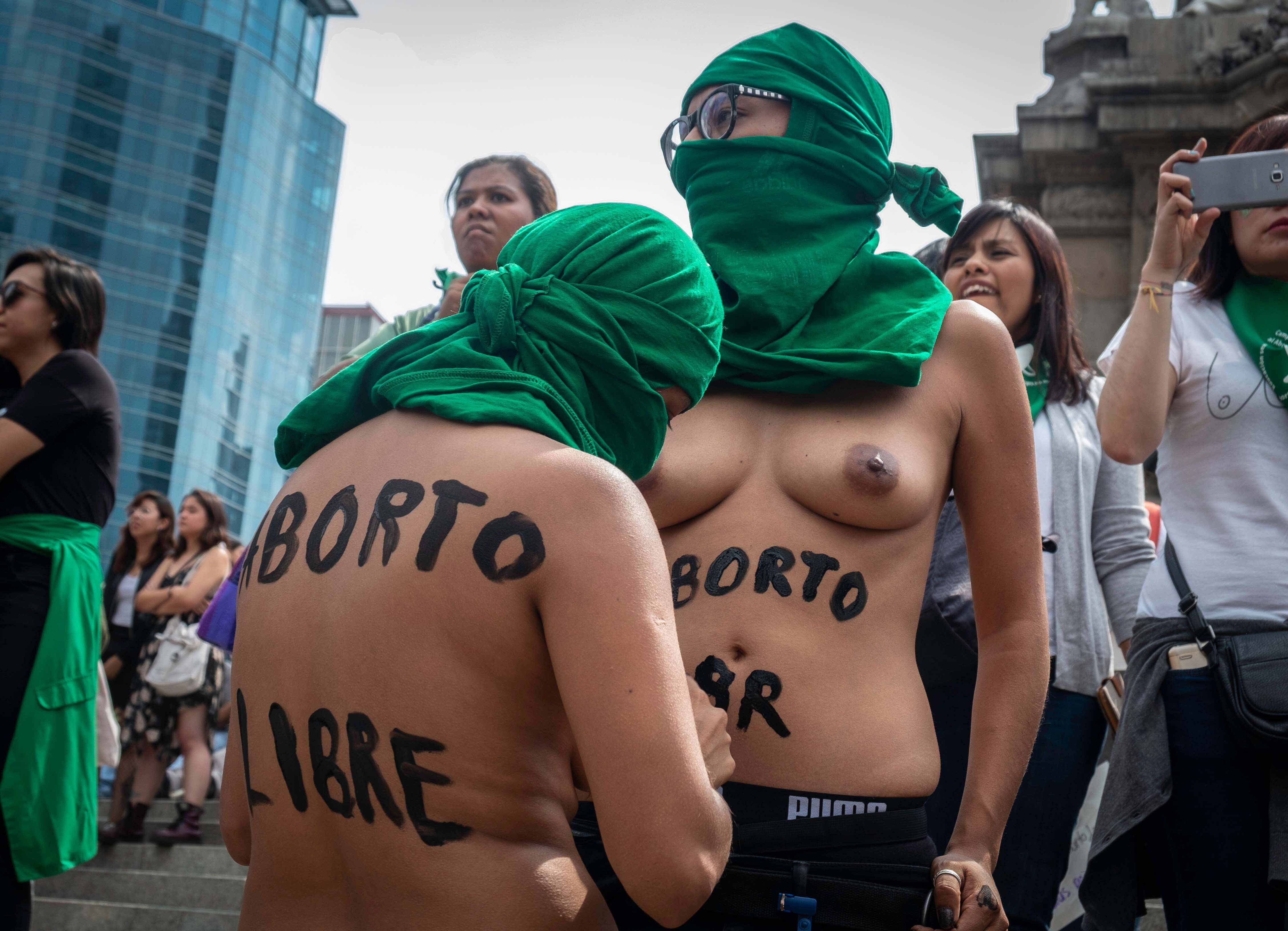 Fotografía en plano medio de dos mujeres con el torso desnudo que utilizan tela verde a modo de pasamontañas. Una de las mujeres tiene escrito en la espalda la frase: "Aborto libre"