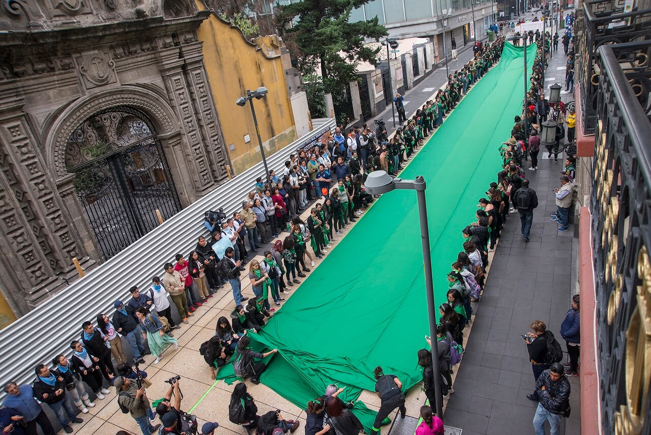 Fotografía en plano general de varias personas que sostienen una tela verde sobre la calle Madero del Centro Histórico de la CDMX.