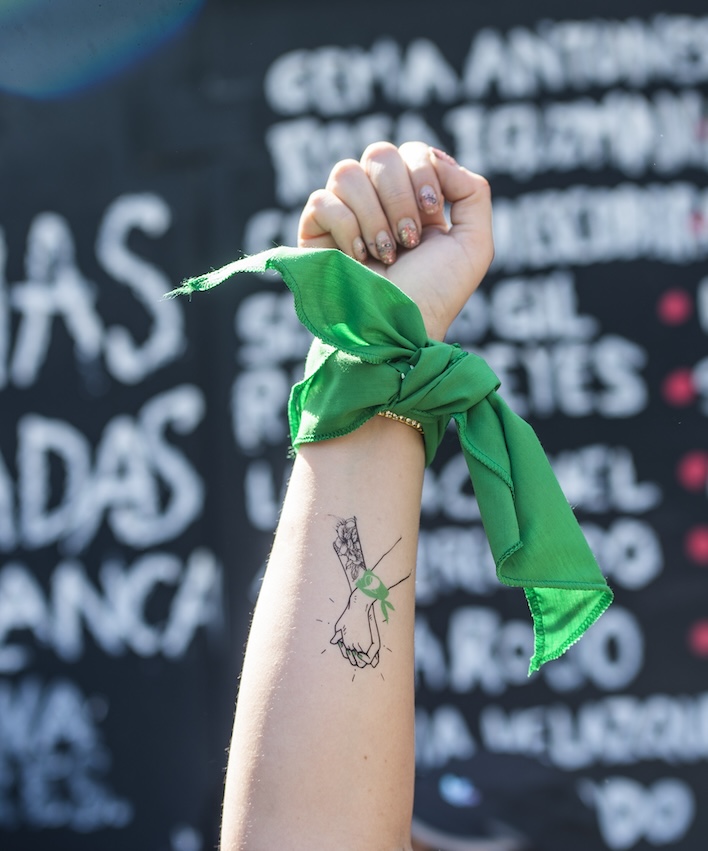 Fotografía en plano detalle de un puño en alto que trae un pañuelo verde abortero.