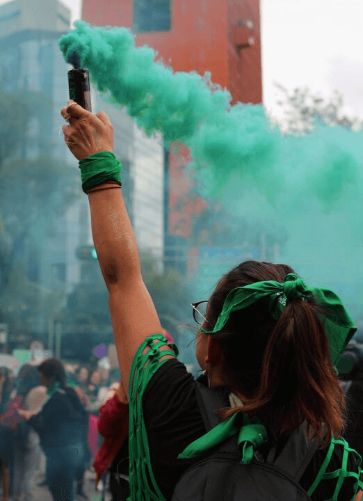 Fotografía en plano medio de una mujer de espaldas que sostiene en su mano una bengala con humo verde durante una manifestación.