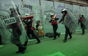 Fotografía en plano general de una mujer tocando el violonchelo en una manifestación. Frente a ella se ven pasar a elementos de la policía femenil.