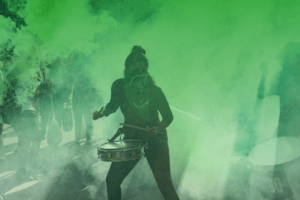 Fotografía en plano medio de una mujer tocando un tambor de batucada durante una manifestación. Tiene el rostro cubierto con un pañuelo verde abortero y está entre humo de una bengala verde.