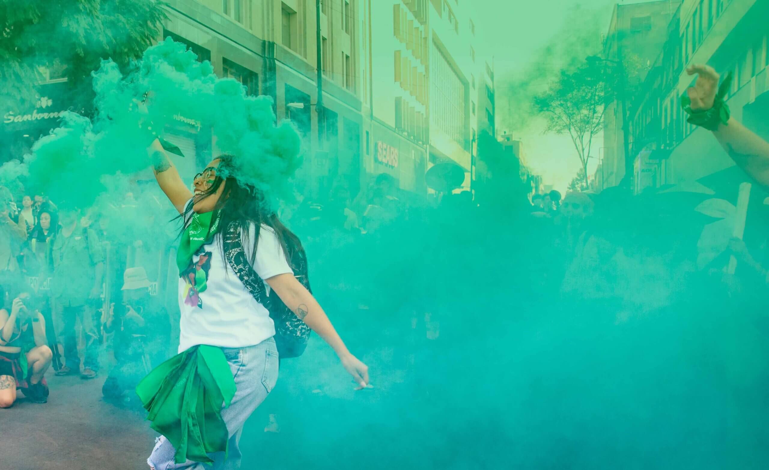 Fotografía en plano medio de una mujer que sostiene en su mano una bengala con humo verde durante una manifestación.